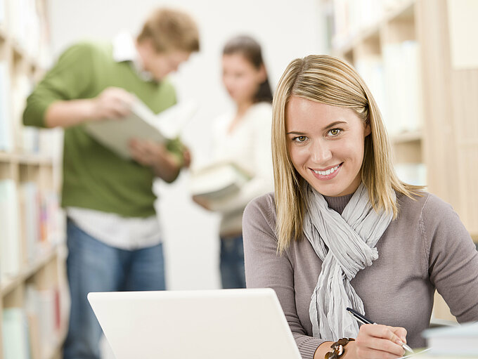kbo-Innovationspreis Auf dem Bild ist eine junge Frau mit ihrem Laptop in einer Bibliothek abgebildet.
